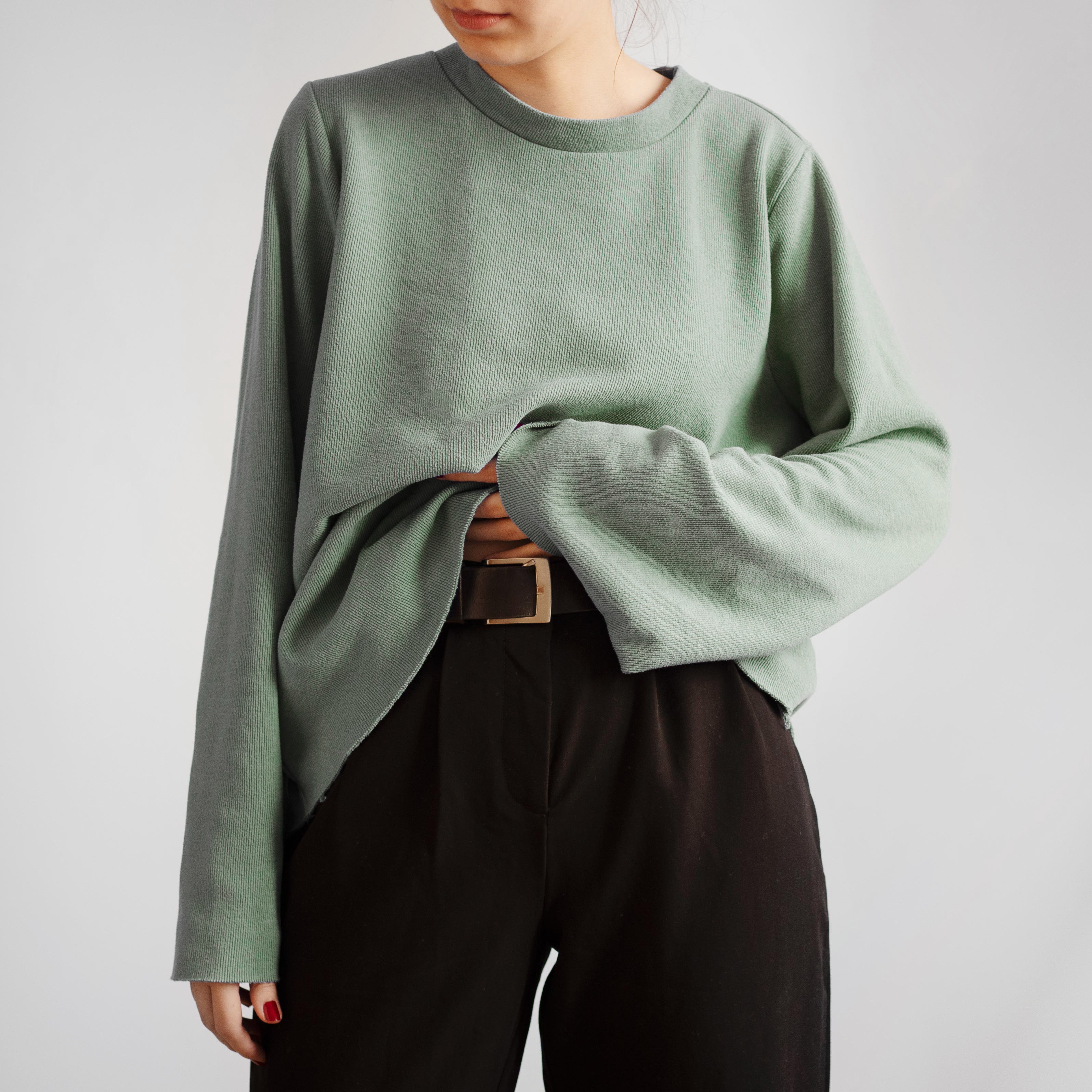 Une femme porte un pantalon noir et un pull vert rentré dans le pantalon pour apercevoir la ceinture