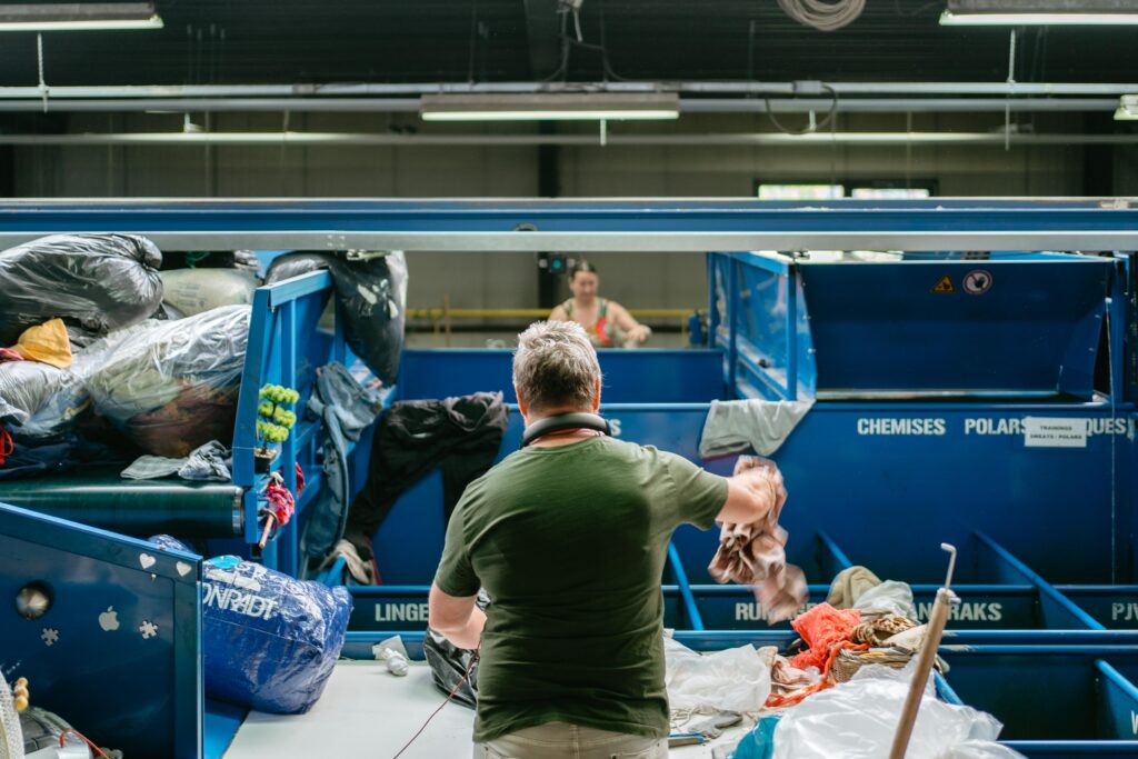 Une personne de dos devant des bacs de tri de vêtements lance un vêtement dans un bac