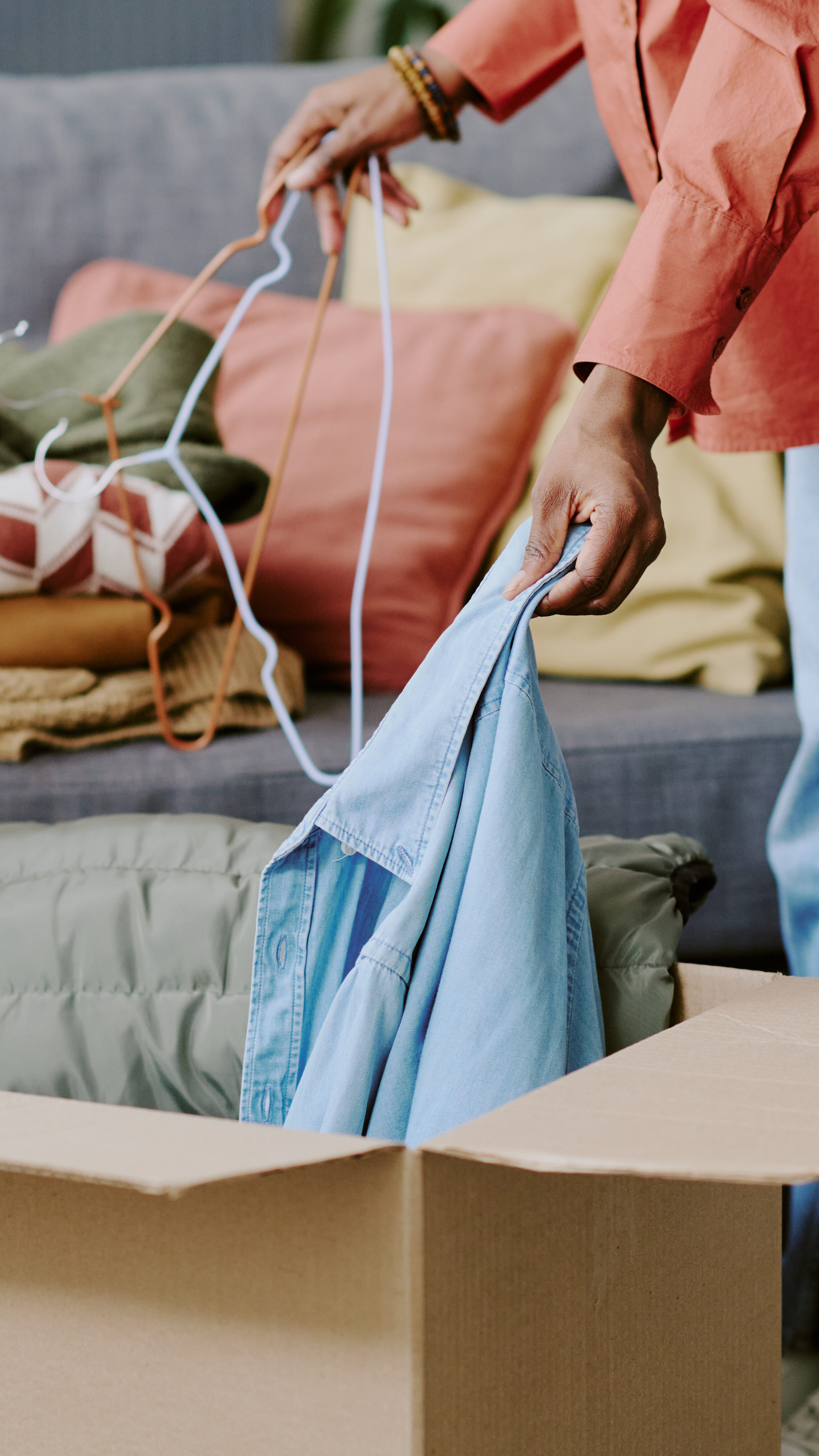 Femme noire rangeant des vêtements pliés dans un carton tout en triant des cintres chez elle, en vue d'un don ou d'un déménagement. Utile pour les associations caritatives, le désencombrement, les déménagements, la revente